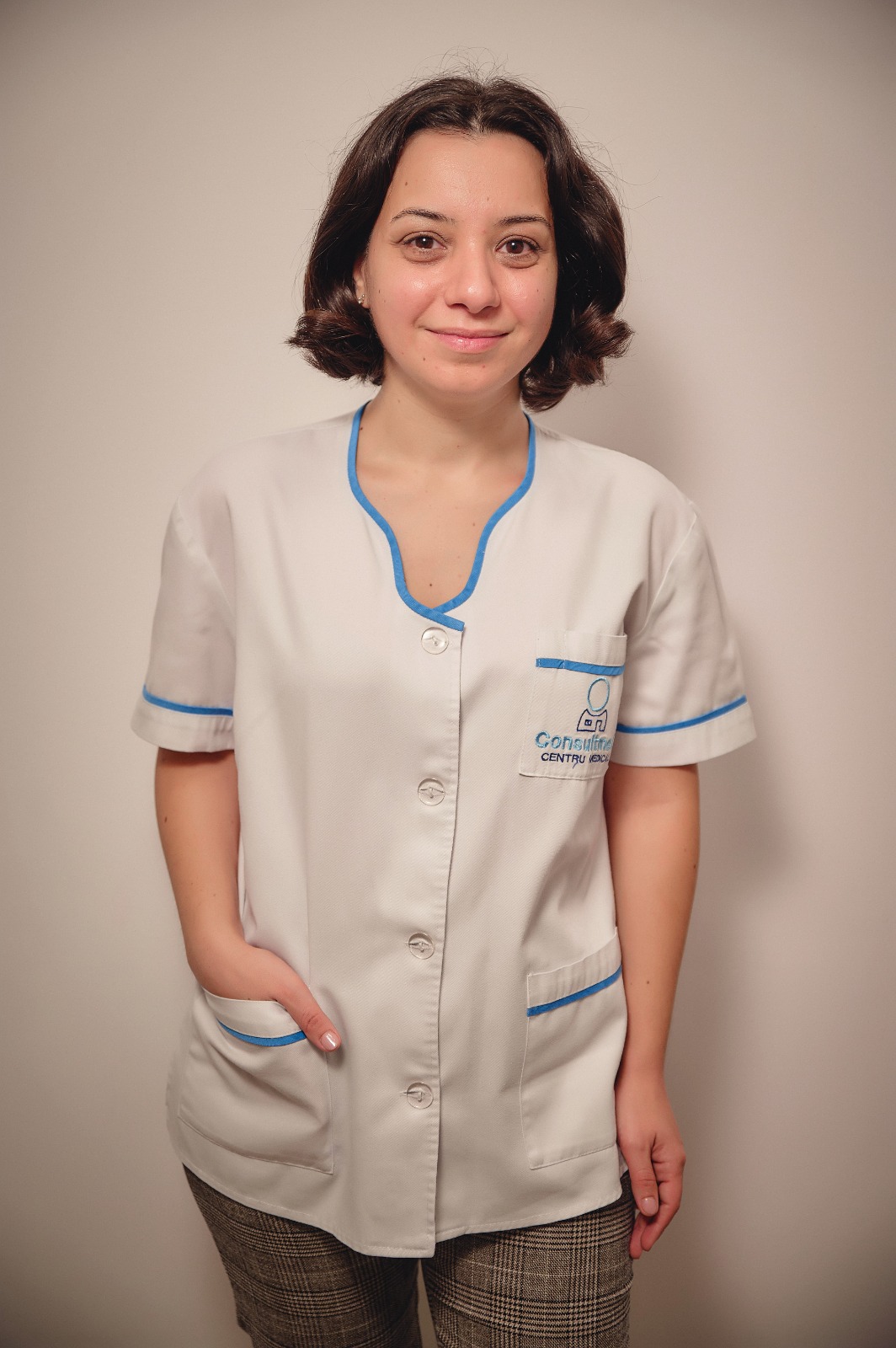 Female healthcare worker in a white uniform with blue piping, standing against a neutral wall and smiling.