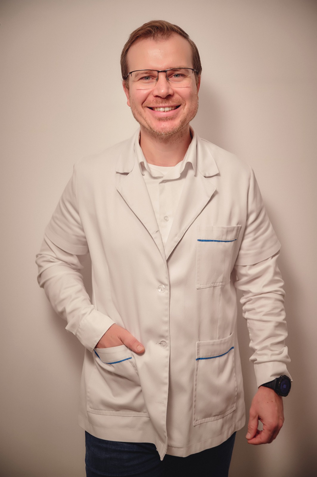 Smiling man wearing a white lab coat and glasses stands against a beige wall.