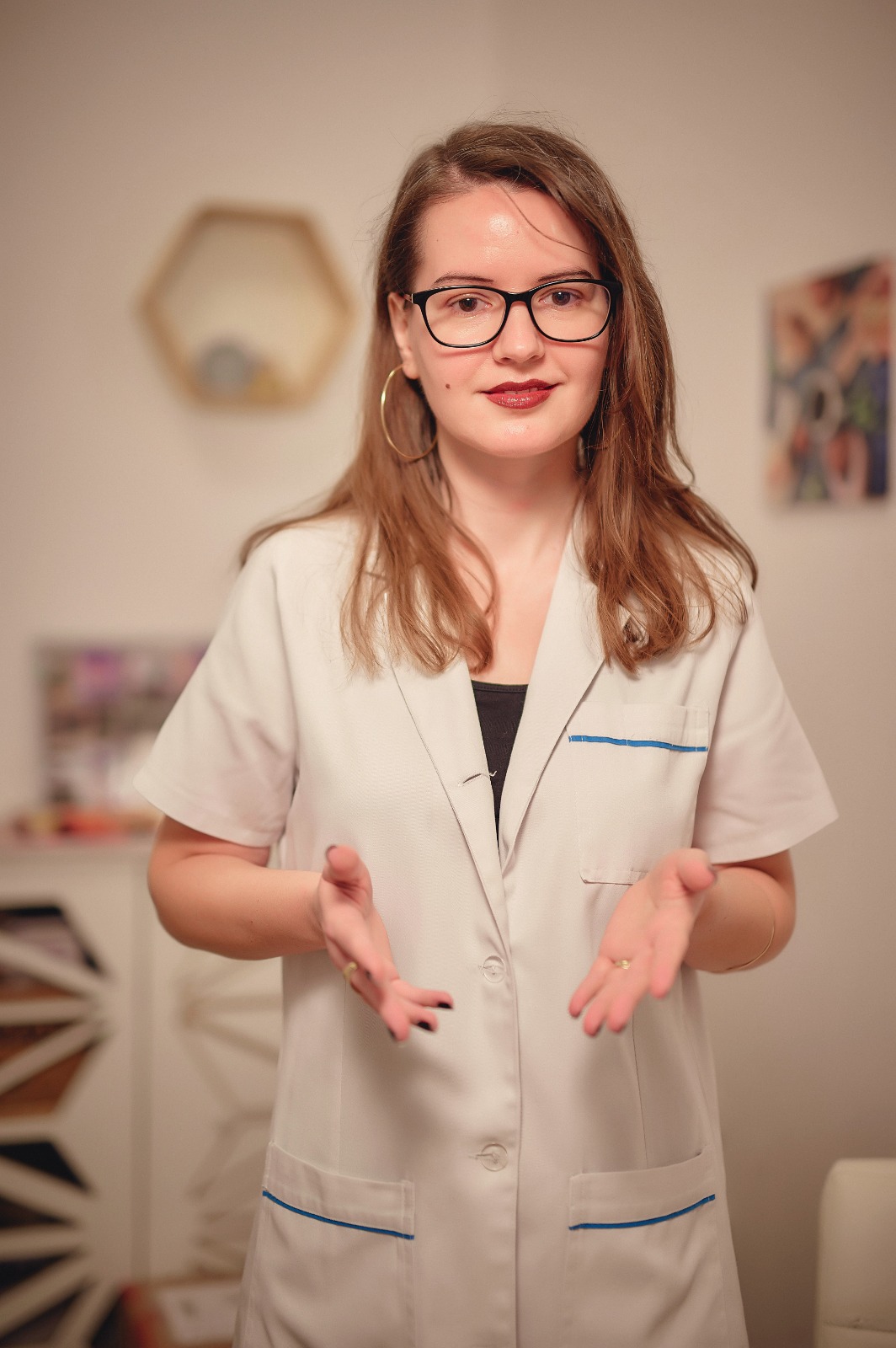 Woman with shoulder-length brown hair and black glasses in a white lab coat, gesturing with hands as she speaks in a cozy room.