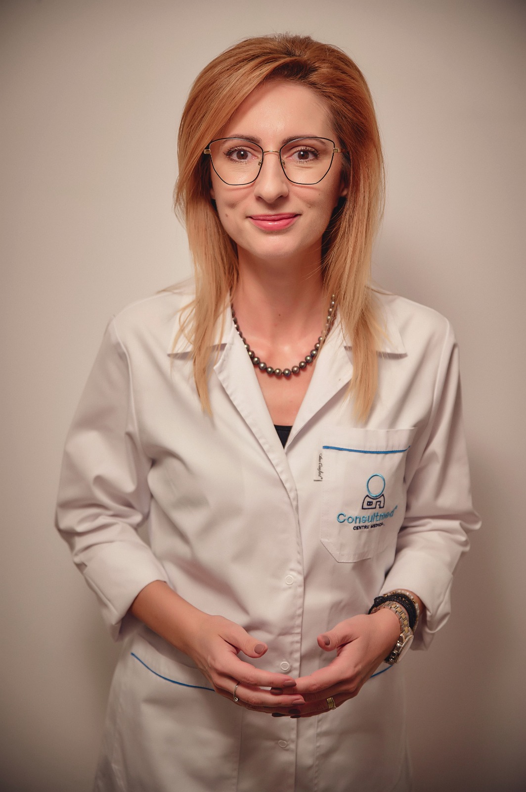 Professional woman with blonde hair and glasses in a white lab coat, standing against a neutral backdrop, hands in front, wearing a beaded necklace and watch.