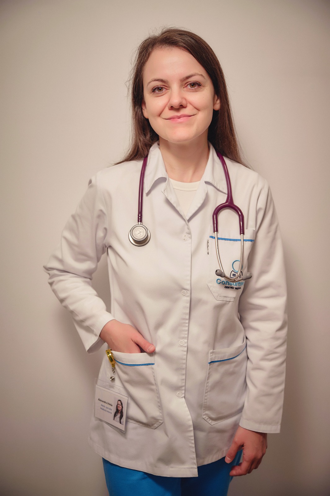 Female clinician in a white coat with stethoscope around her neck, smiling at the camera against a plain wall, wearing an ID badge.