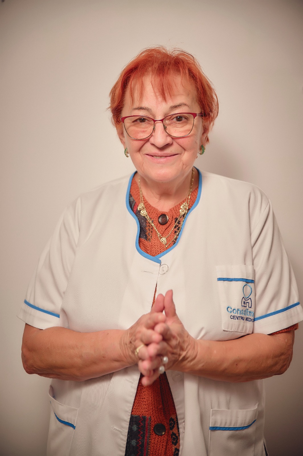 Older female healthcare professional in a white coat with blue trim, hands clasped, smiling at the camera.