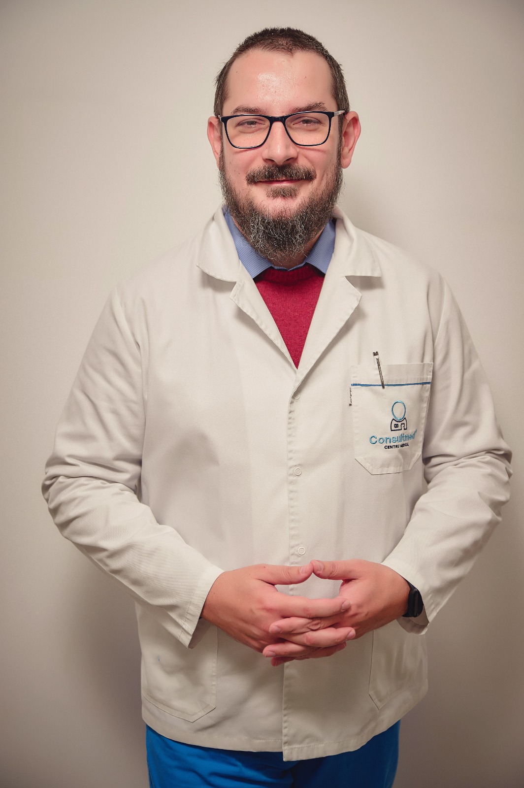 Male healthcare professional wearing a white lab coat over a blue shirt and red sweater, with glasses, standing against a neutral background.