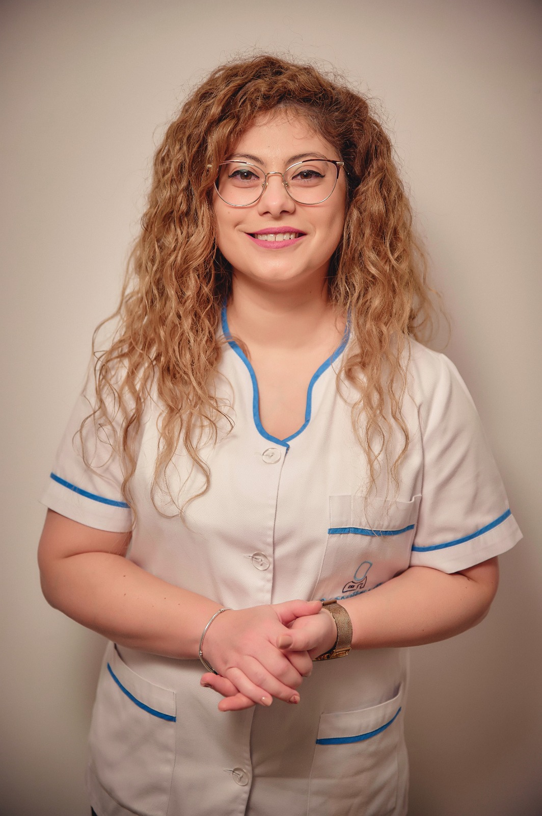 Young female healthcare worker with curly hair and glasses, in a white medical uniform with blue trim, smiling at the camera.