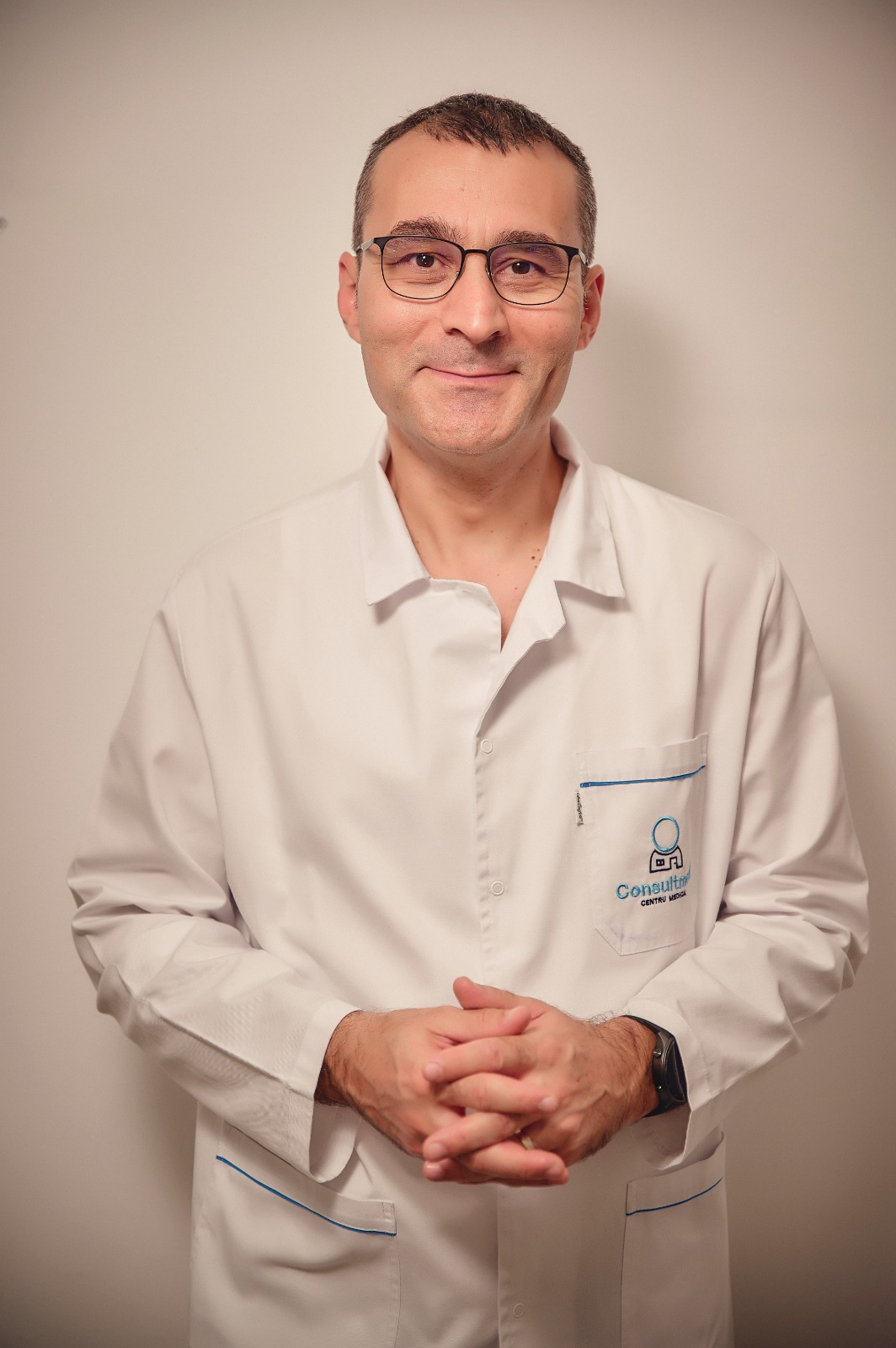 Portrait of a man in a white medical coat with glasses, smiling gently, arms lightly folded.